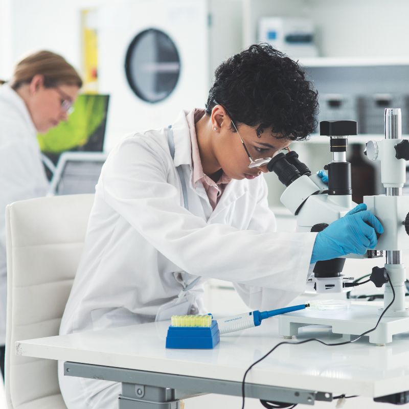 A pair of biotech researchers in a lab, with one looking at a formula under the microscope.